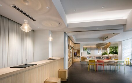 A modern interior with a wooden reception desk, a dining area with colorful chairs, and a kitchen with plants.