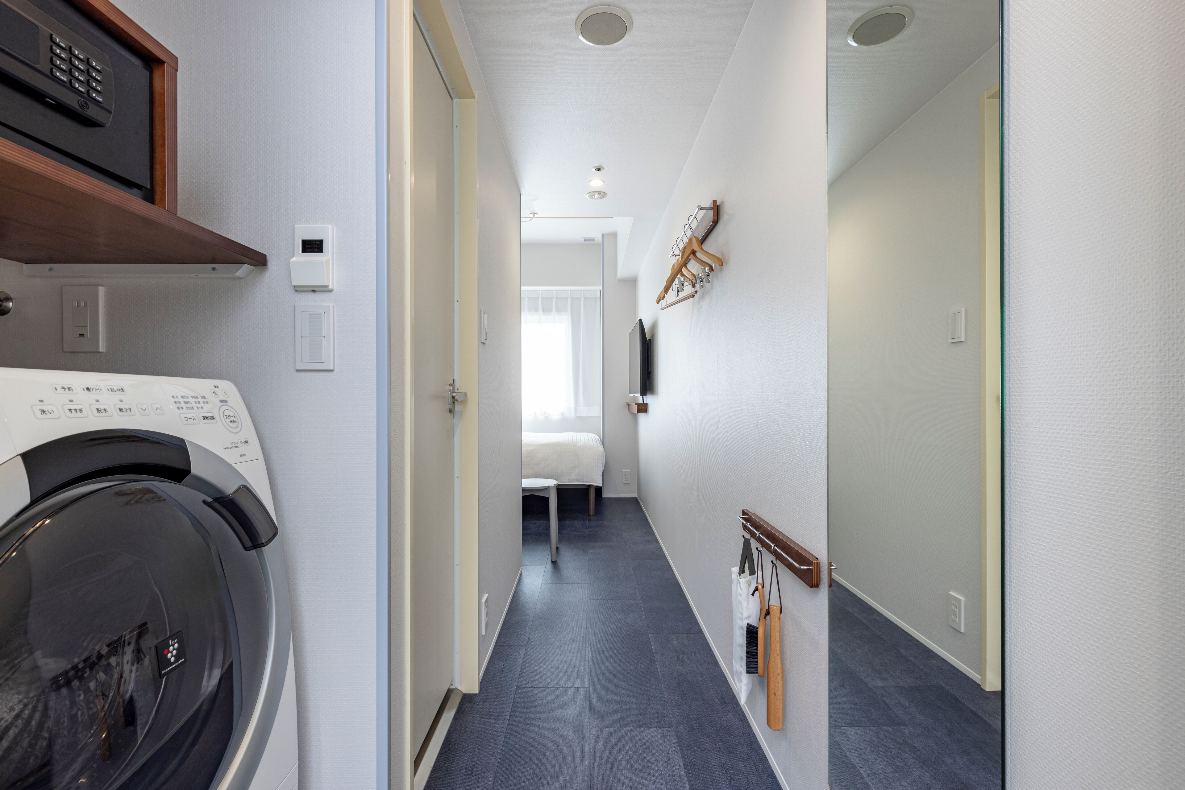 A narrow hallway with a washing machine on the left, a bed in the background, and a coat rack on the right.