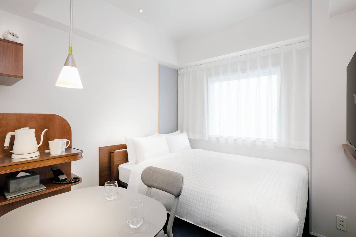A hotel room with a bed, a bedside table with a kettle and cups, a chair, a round table with a glass, and a window with curtains.