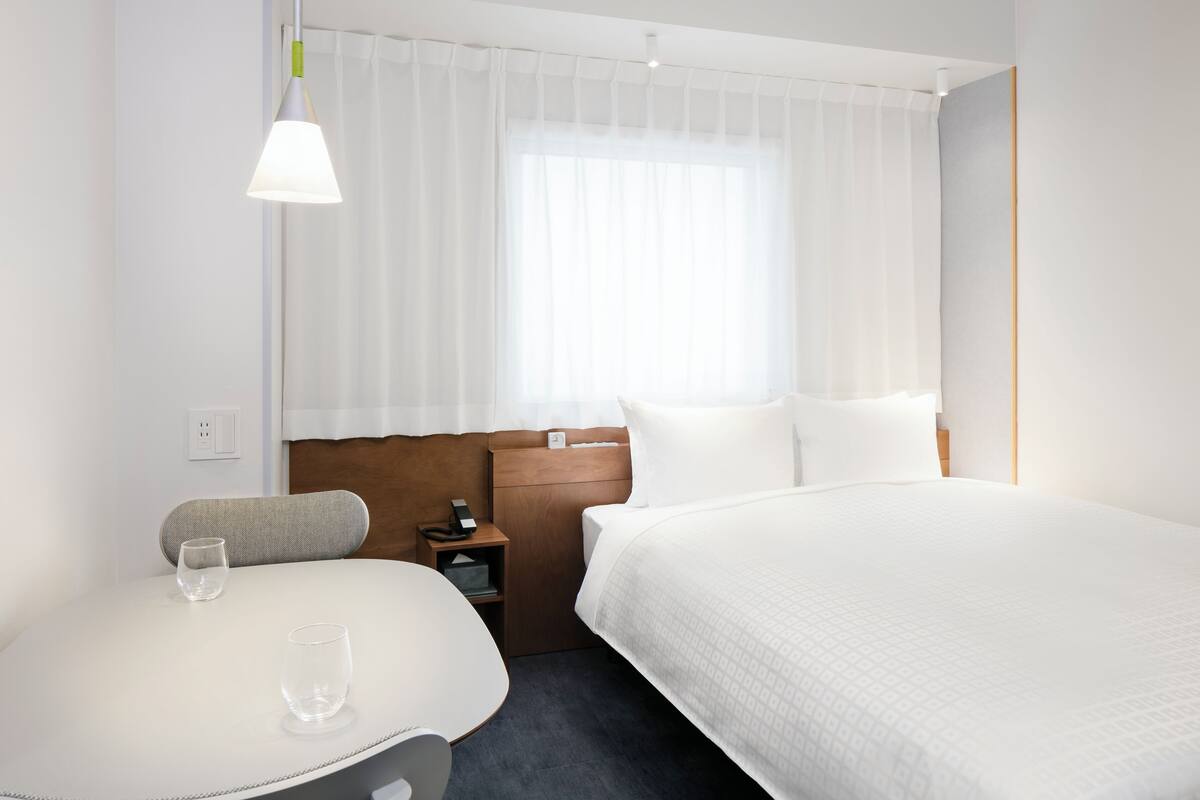 A hotel room with a bed, a bedside table with a lamp, a chair, and a window with curtains.