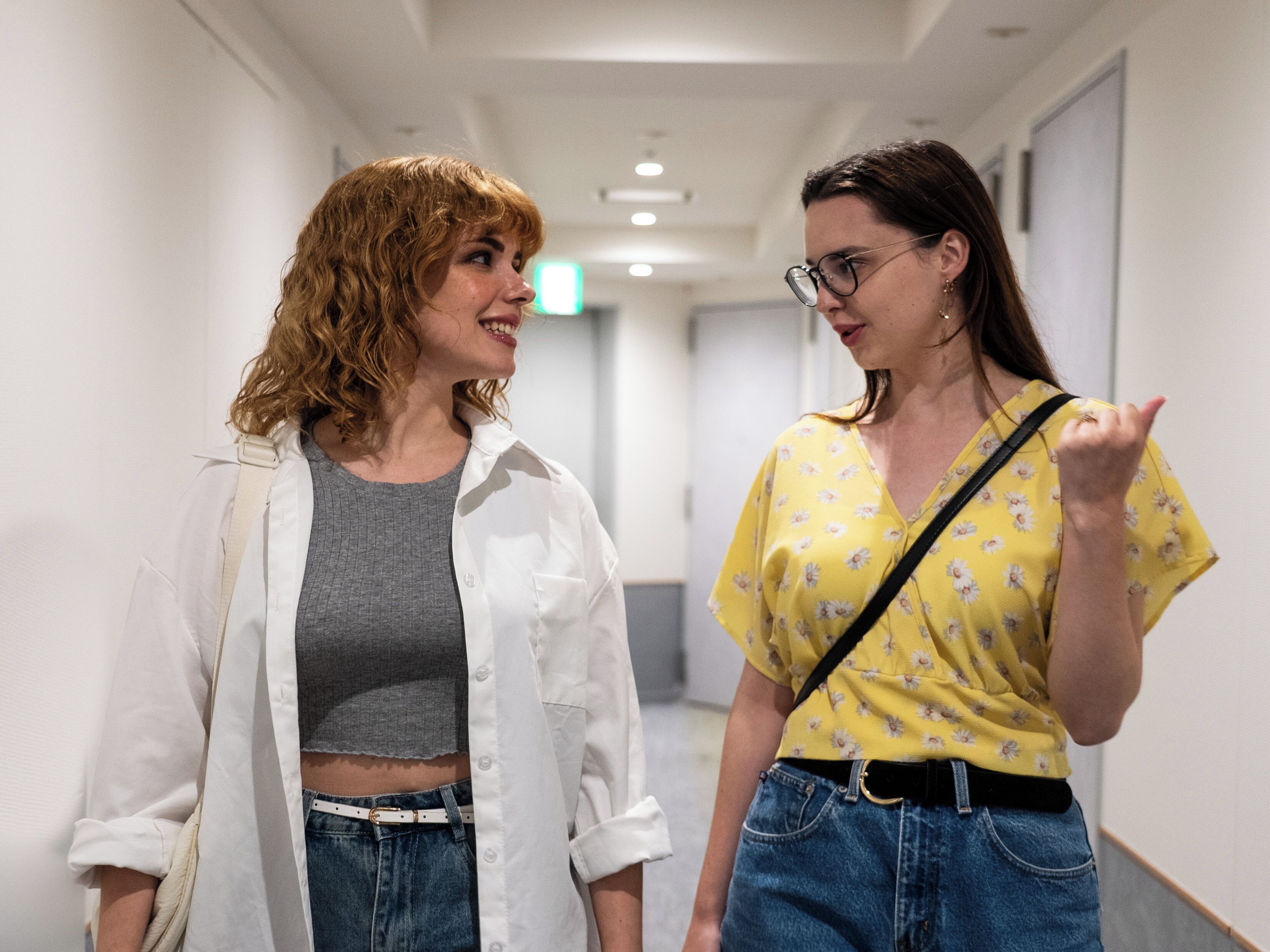 Two women walking down a hallway with white walls and a ceiling with recessed lighting.