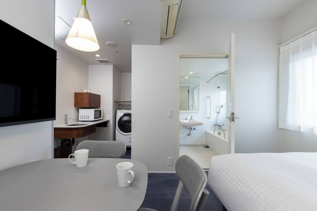 A hotel room with a bed, a table with two white mugs, a microwave, a washing machine, and a bathroom visible through an open door.