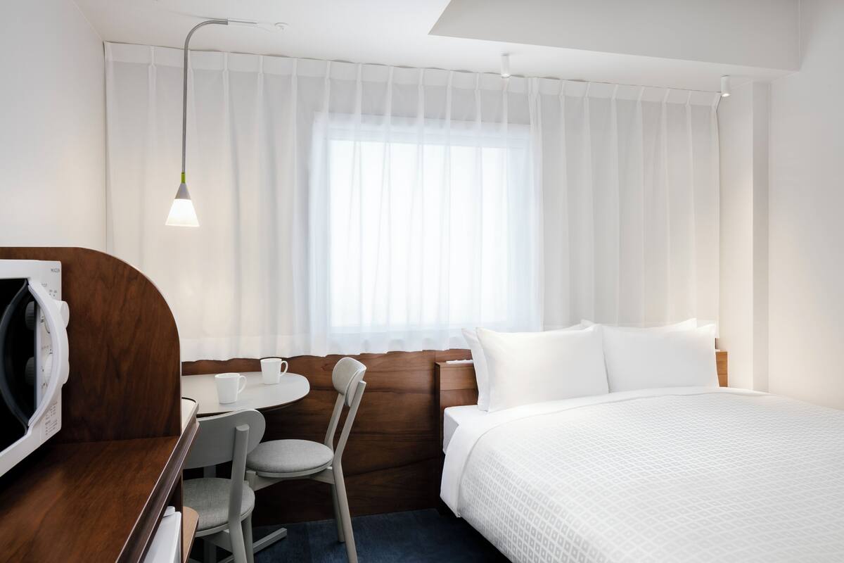 A hotel room with a bed, a desk with a chair, a microwave, and a window with sheer curtains.