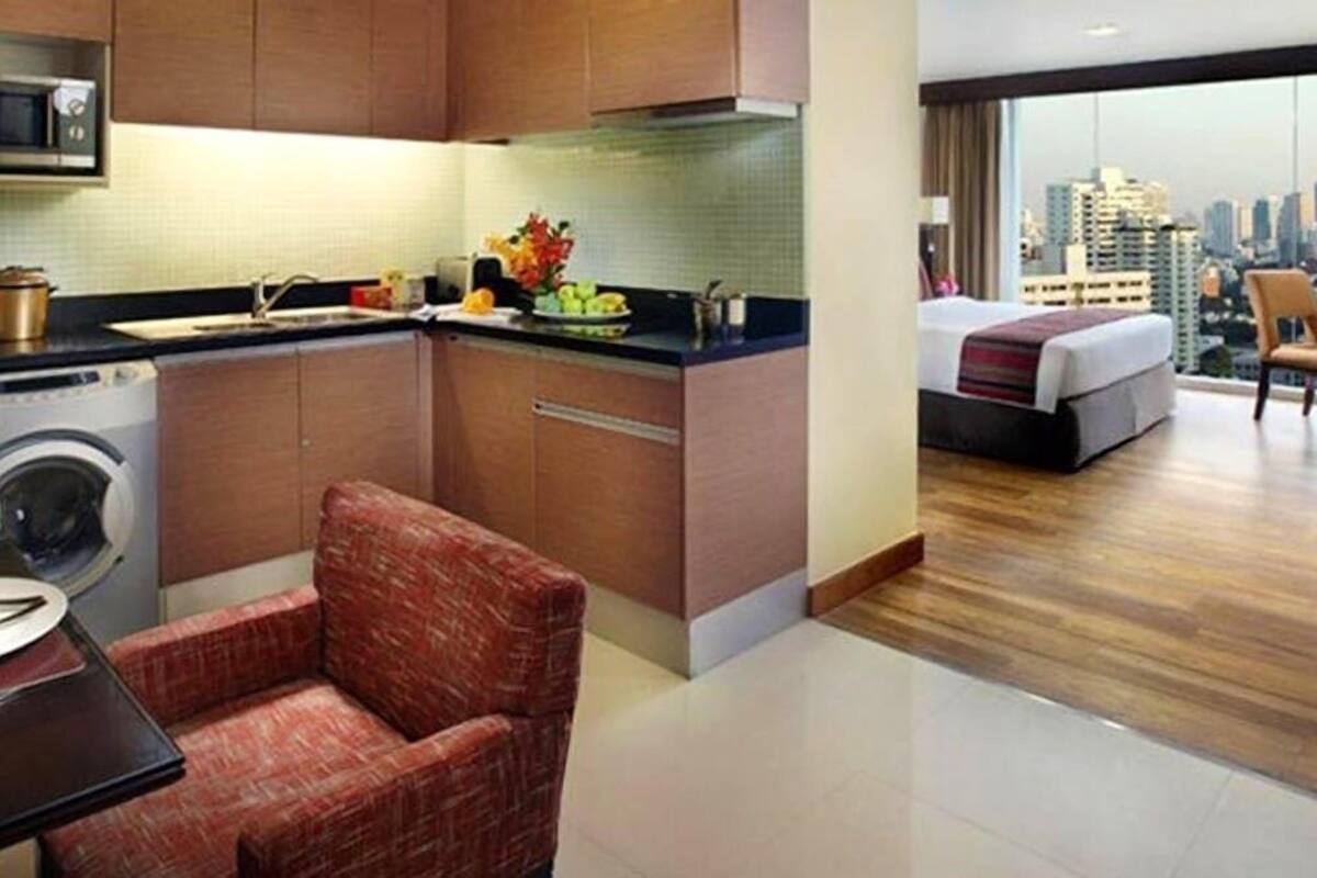A hotel room with a kitchen area, a washing machine, a dining table, and a bed with a city view.