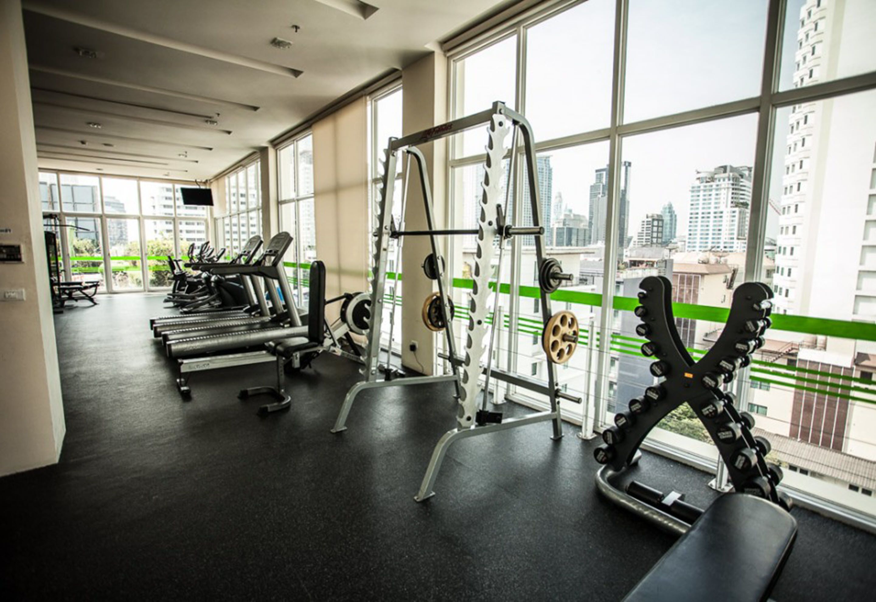 A gym with treadmills, weight machines, and a view of a cityscape through large windows.