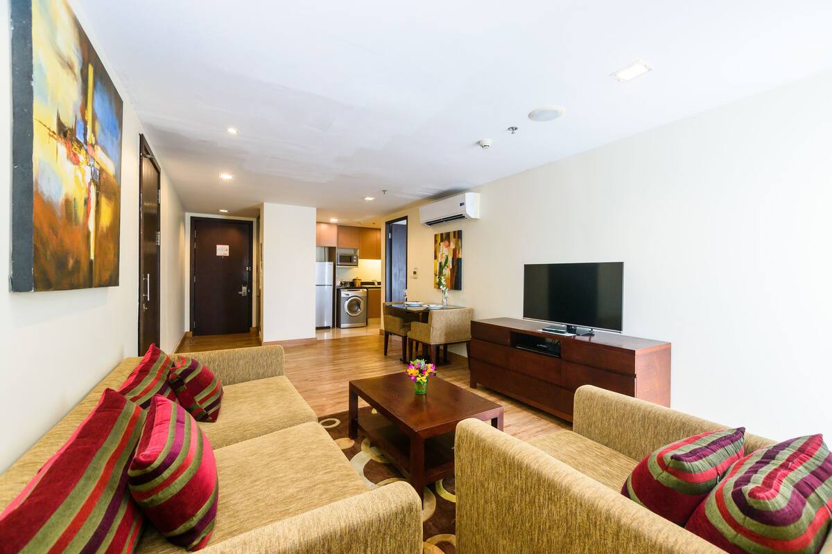 A living room with a sofa set, a coffee table, and a television. There is a kitchen area visible in the background.