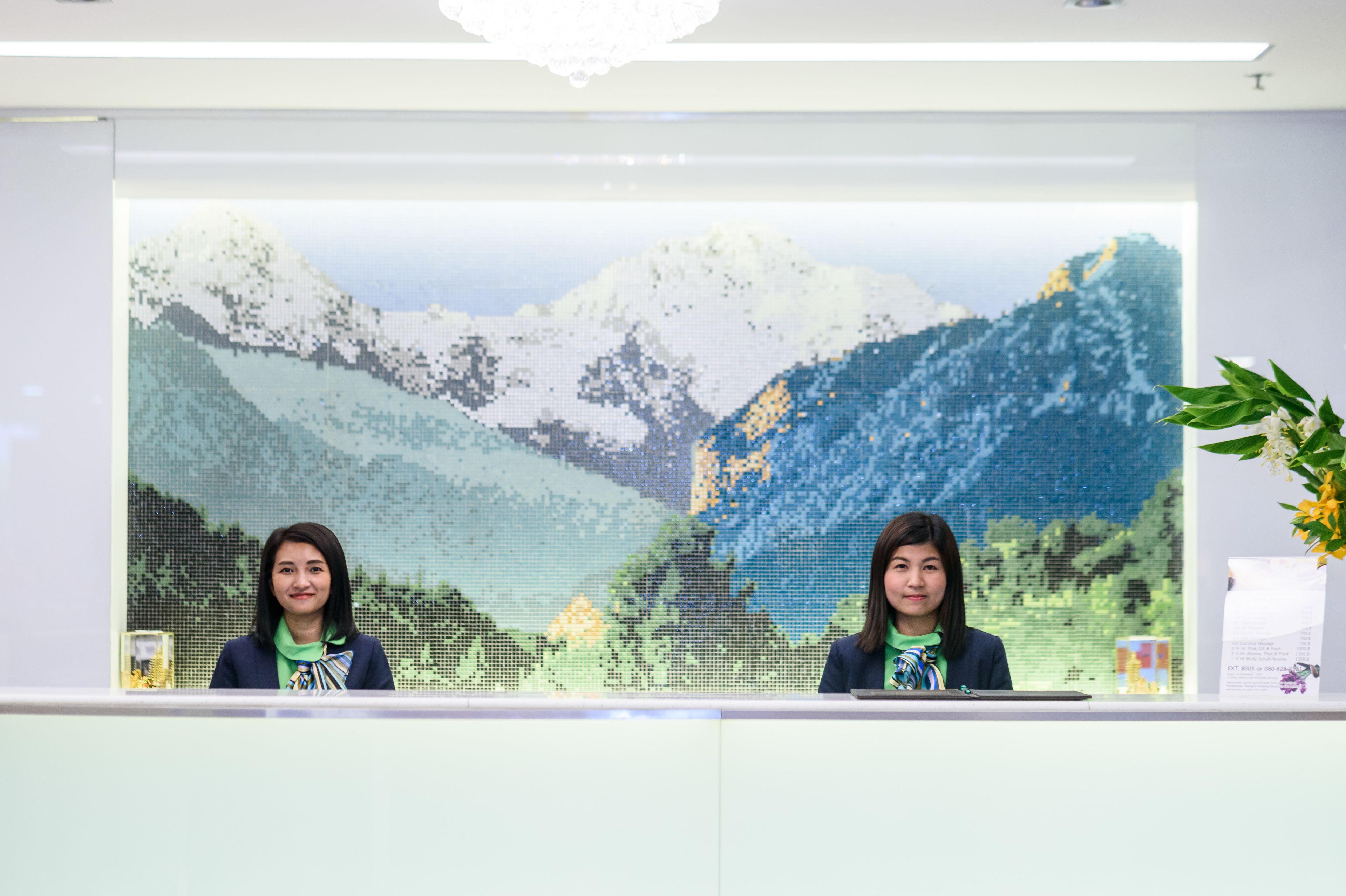 Two receptionists at a desk with a large mosaic artwork in the background.