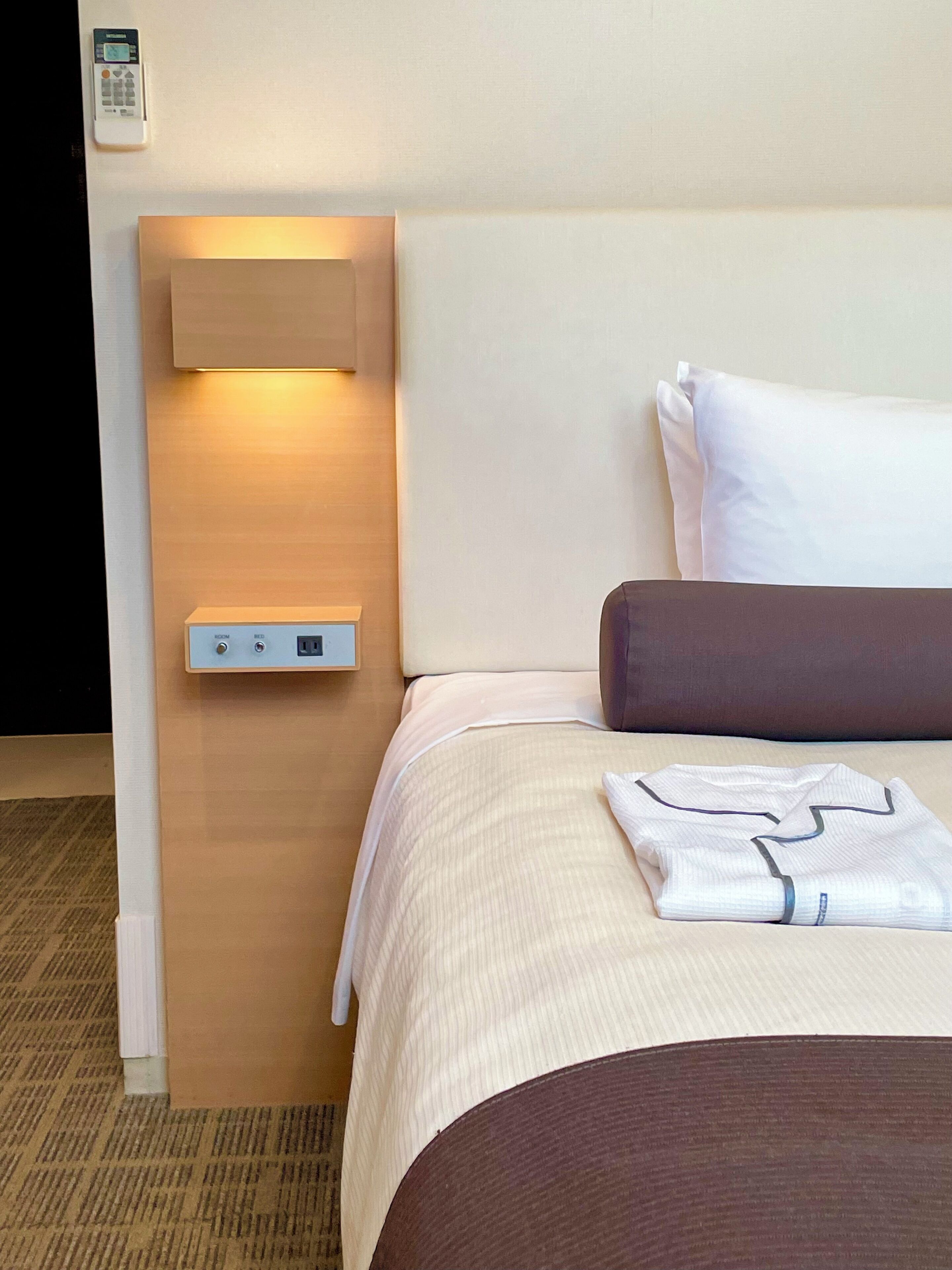 A neatly made bed with a white and brown comforter, a folded white shirt on the bed, and a wall-mounted control panel with a power outlet.