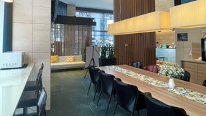A modern hotel lobby with a long wooden reception desk, black leather chairs, a sofa with floral cushions, a large potted plant, and a view of the street through large windows. - Tokyu Stay Kamata - Tokyo Haneda (Tokyo)