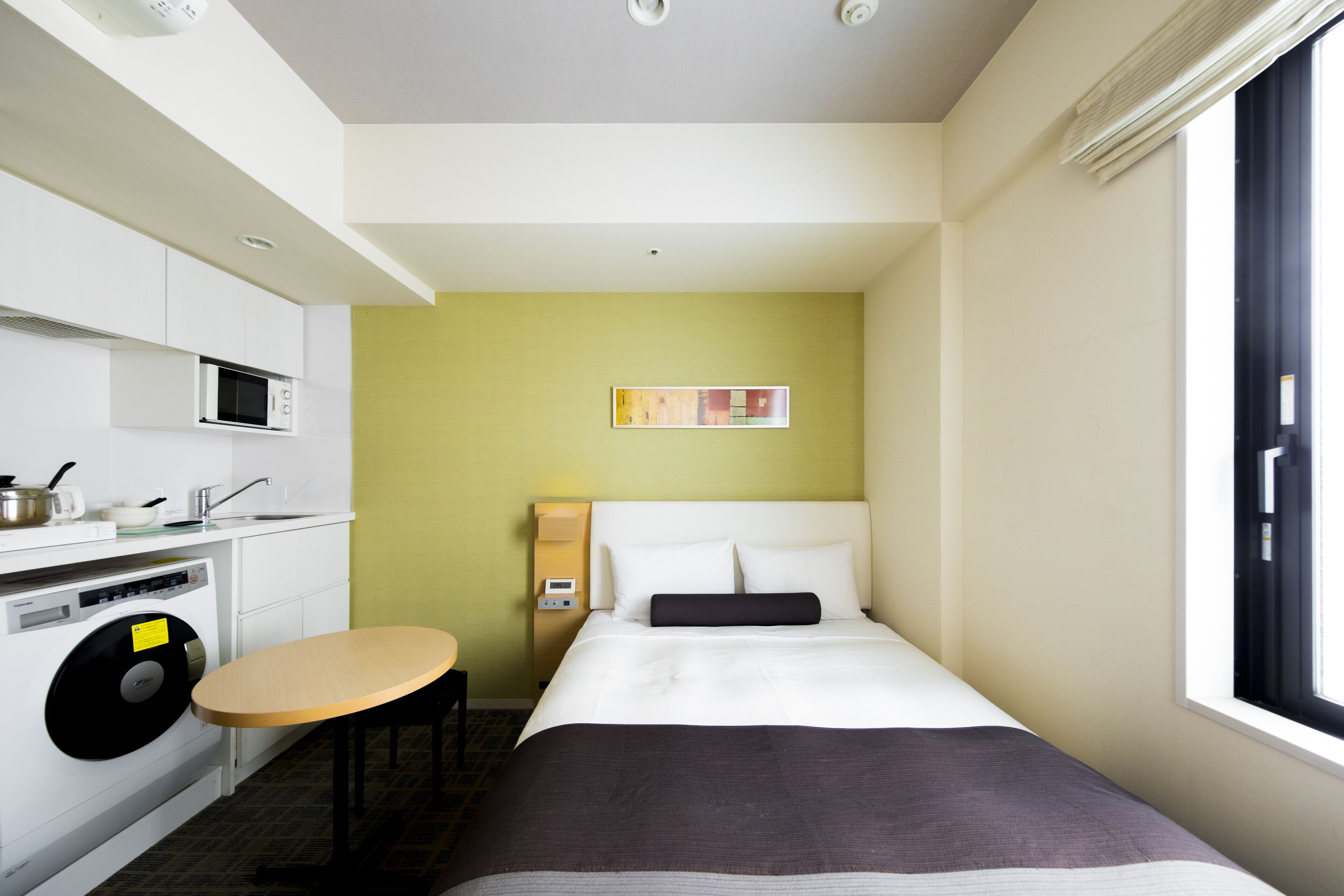 A hotel room with a bed, a small table, a washing machine, a microwave, and a window with blinds.