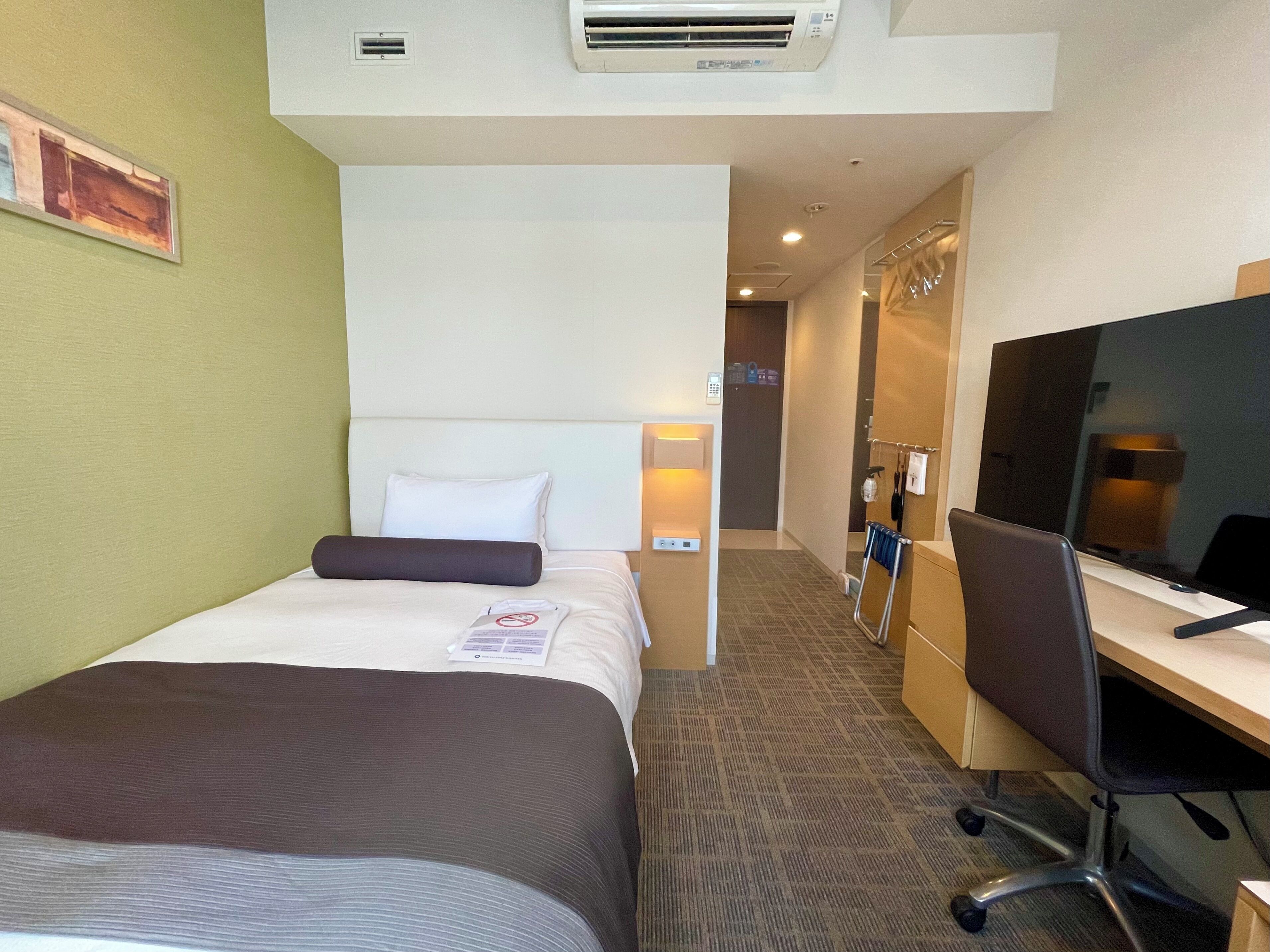 A hotel room with a bed, a desk, a chair, a TV, and a wall-mounted air conditioner.