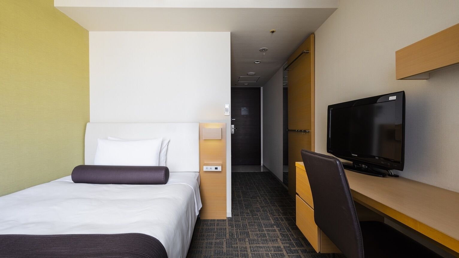 Une chambre d’hôtel avec un lit, un bureau avec une télévision et une porte donnant sur un couloir.