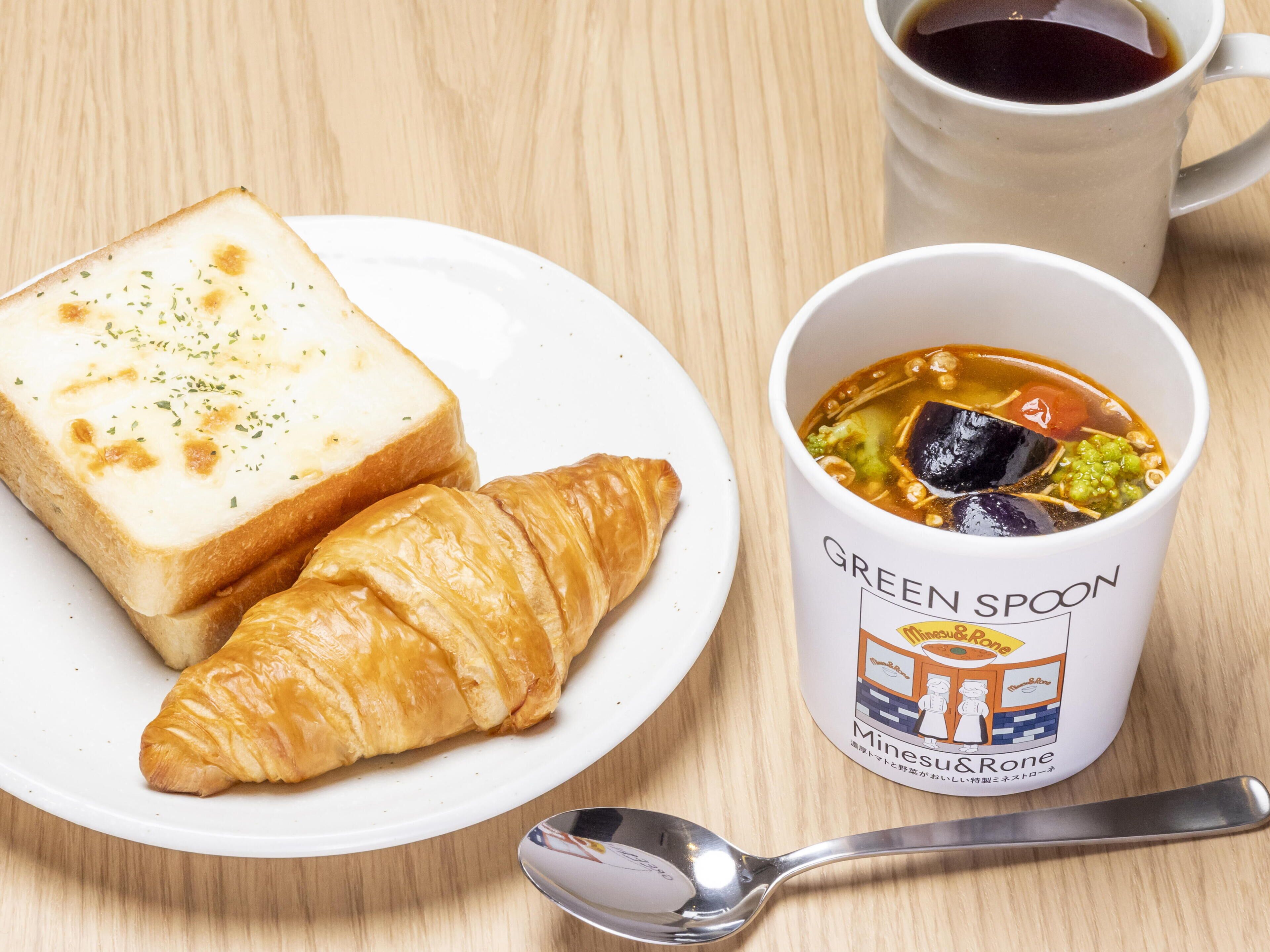 Lautasella on viipale leipää ja croissant, mukissa lukee ”GREEN SPOON Mines & Rone”, ja sen vieressä on lusikka.