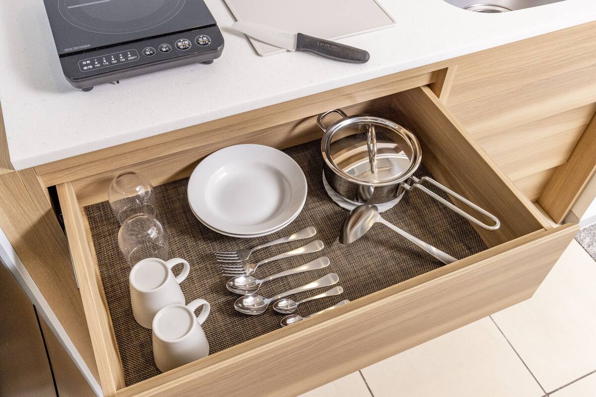 A drawer with kitchen utensils, including a pot strainer, spoons, forks, and a white plate.
