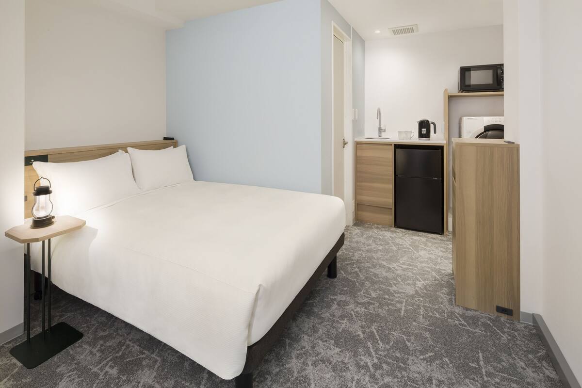A hotel room with a bed, bedside table with a lamp, a small kitchenette with a microwave, and a refrigerator.