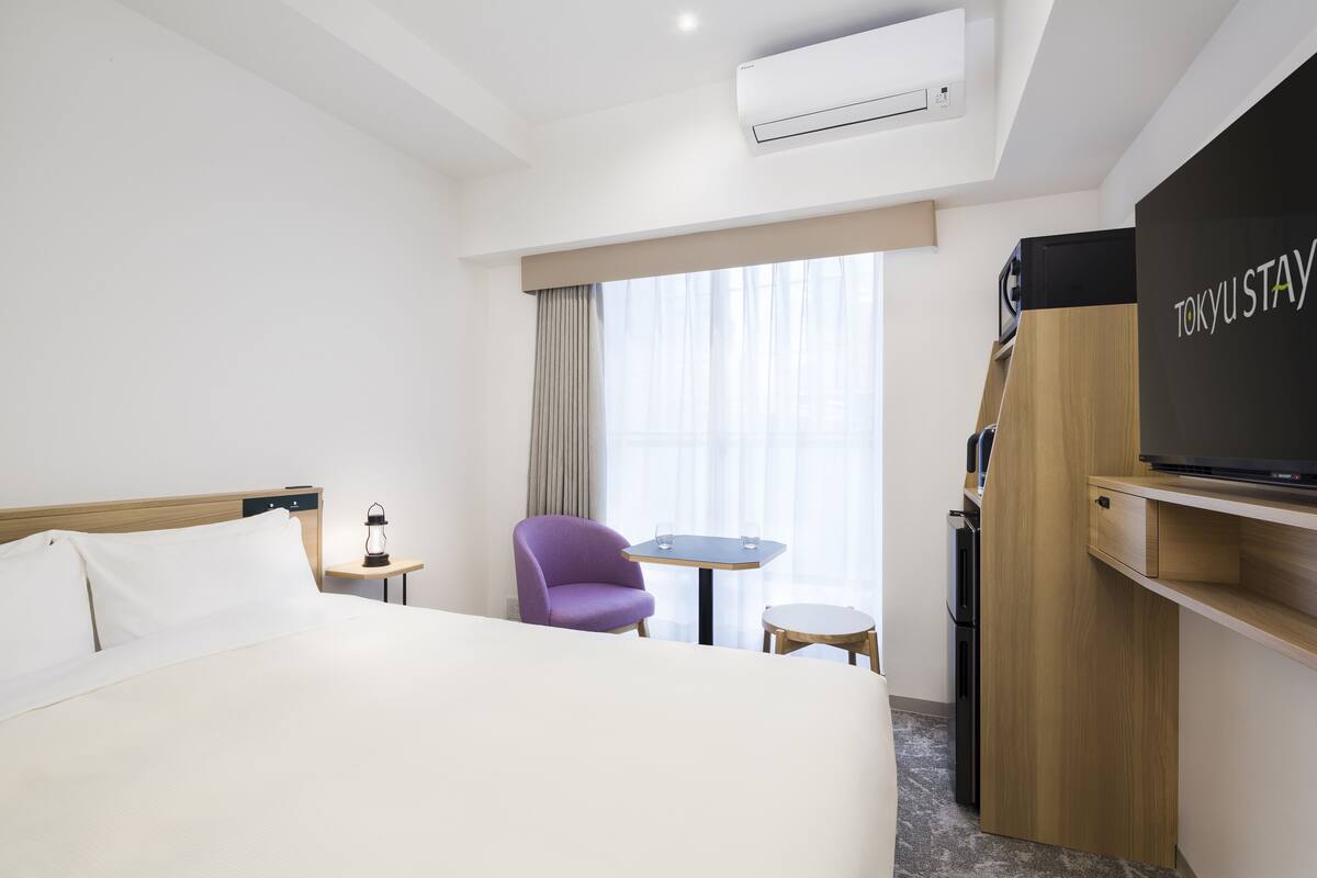A hotel room with a bed, a purple armchair, a small table, a TV, and a minibar.