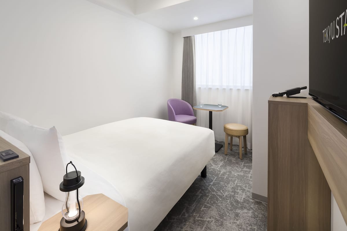 A hotel room with a bed, a small table with two glasses, a chair, a TV, and a lamp.