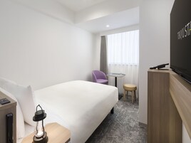 A hotel room with a bed, a small table with two glasses, a chair, a TV, and a lamp.