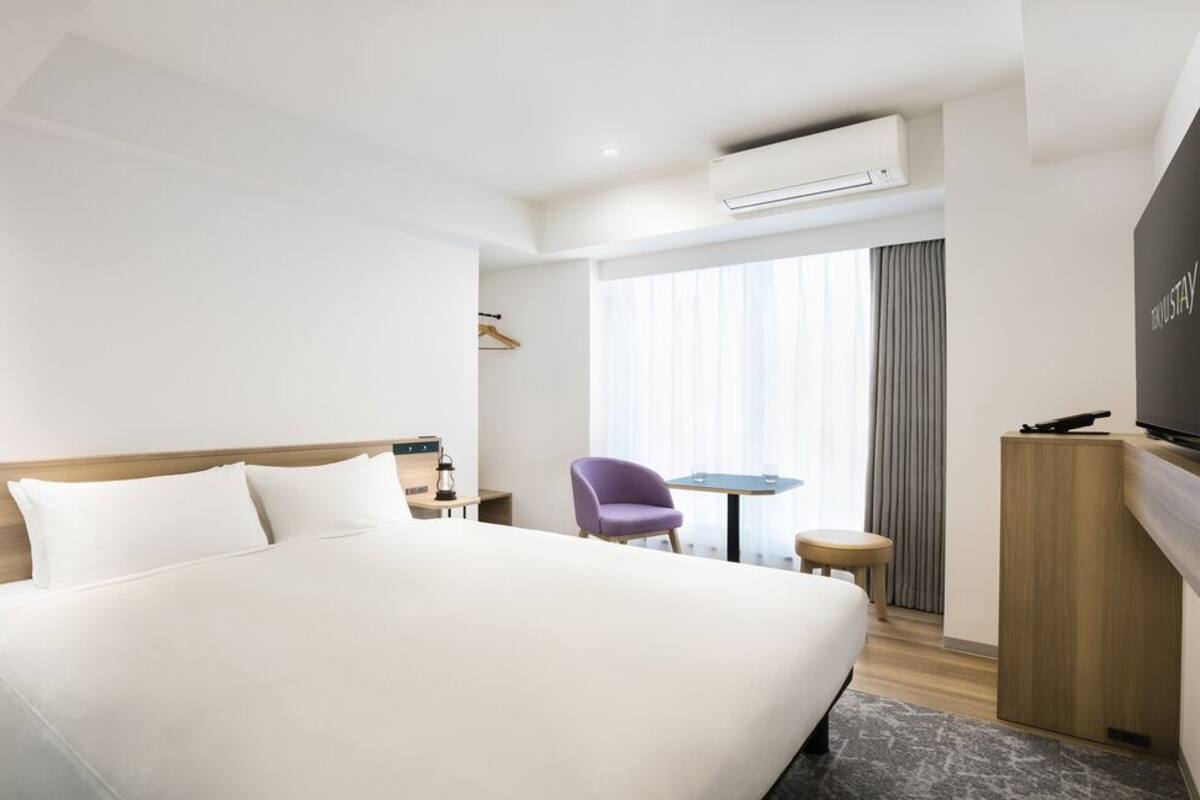 A hotel room with a large bed, a TV, a small table, a chair, and an air conditioning unit.