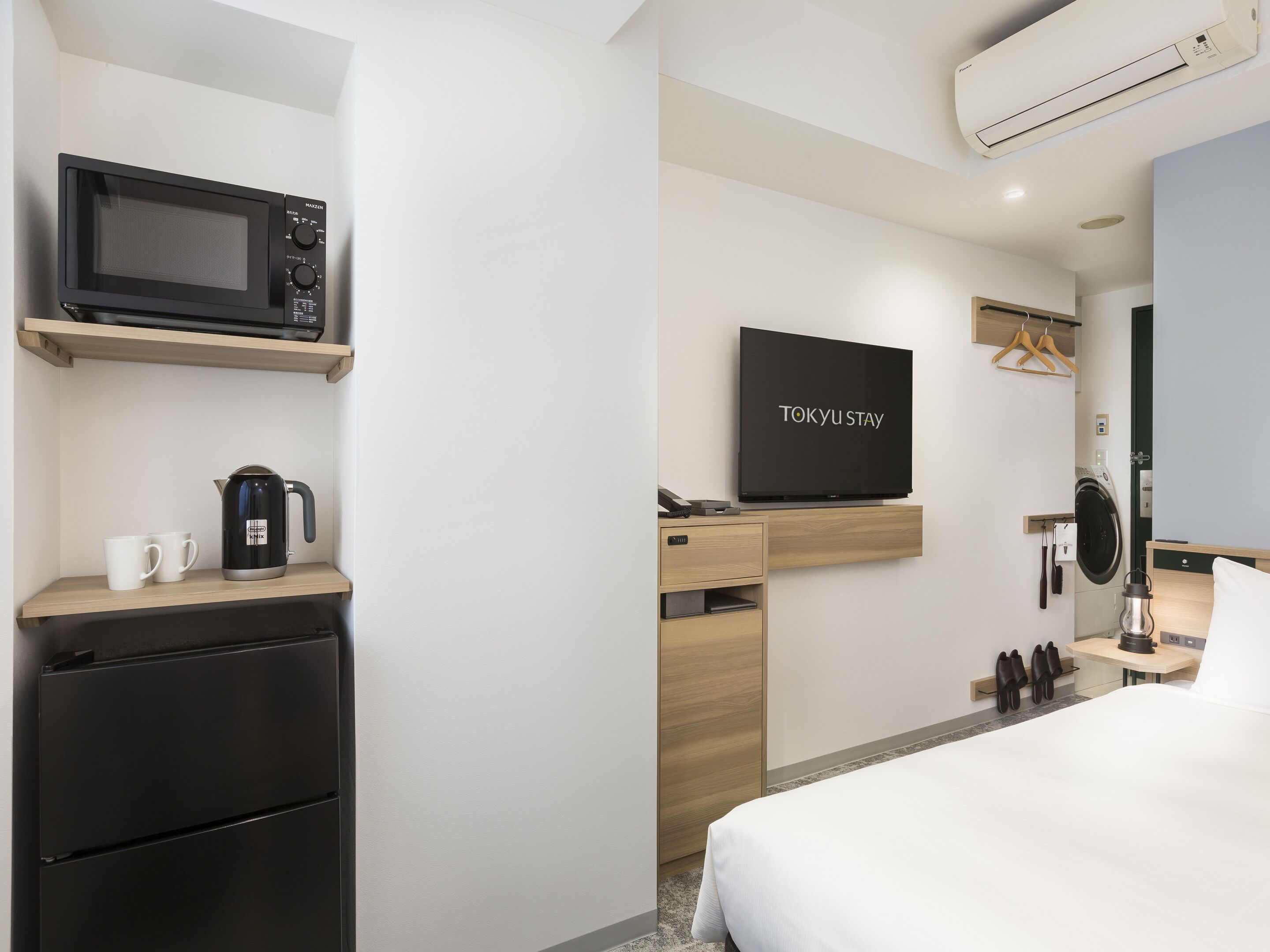 A modern hotel room with a bed, a flat-screen TV, a microwave, a kettle, a mini-fridge, a desk, and a hanging shower curtain.