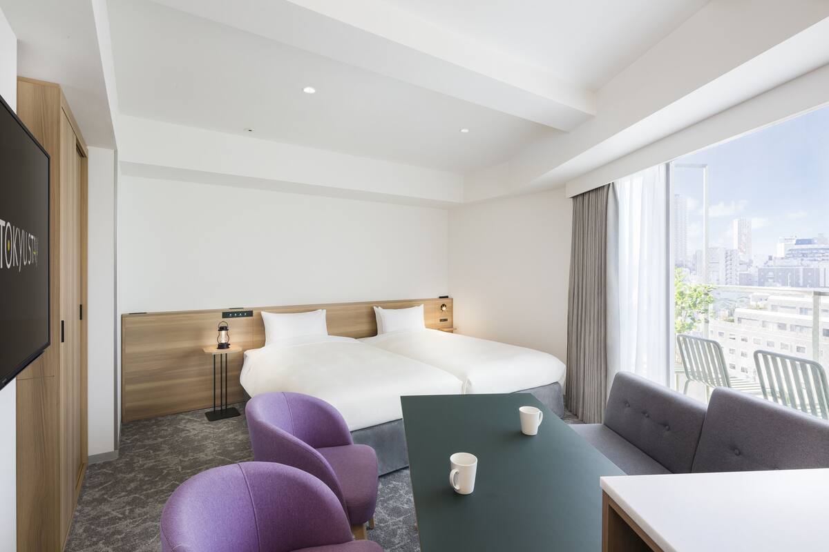 A hotel room with a large bed, a desk, purple chairs, a green table, and a city view.