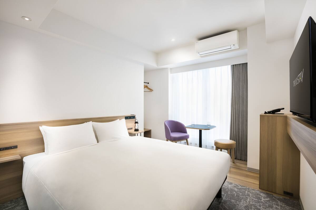 A hotel room with a large bed, a TV, a small table, a chair, and an air conditioning unit.