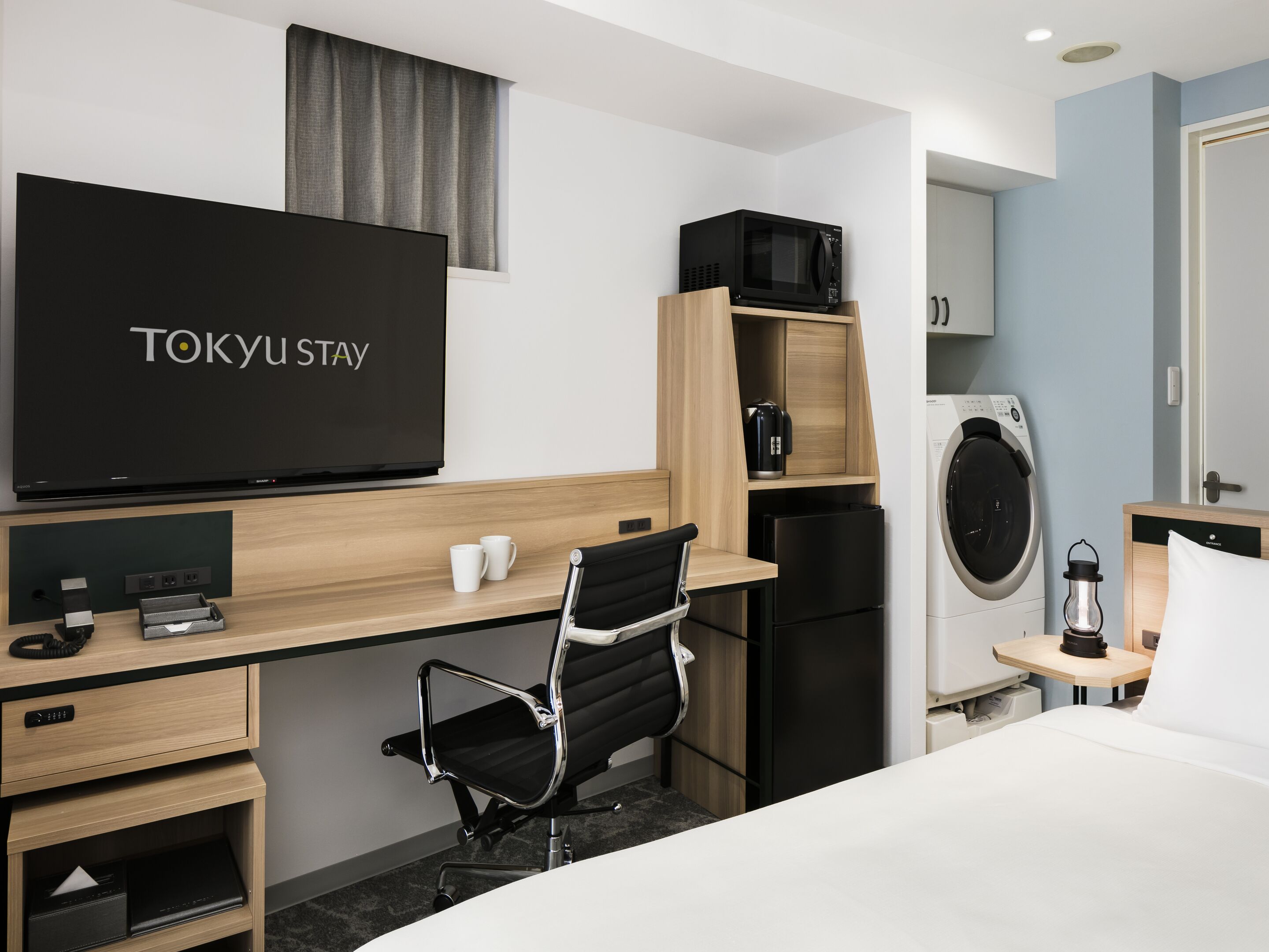A hotel room with a bed, a desk with a computer, a chair, a TV, a microwave, a refrigerator, a washing machine, and a bedside table with a lamp.