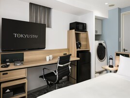 Ein Hotelzimmer mit einem Bett, einem Schreibtisch mit Computer, einem Sessel, einem Fernseher, einer Mikrowelle, einem Kühlschrank, einer Waschmaschine und einem Nachttisch mit Lampe.