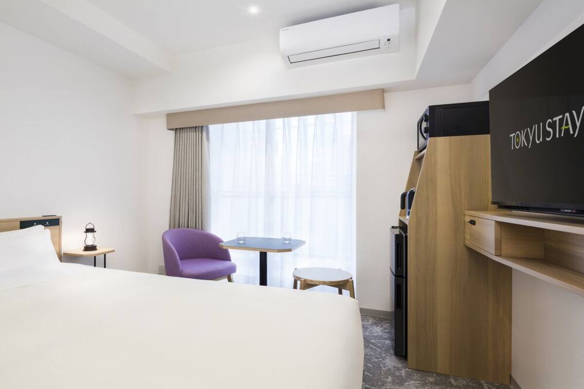 A hotel room with a bed, a TV, a chair, a small table, and a window with curtains.