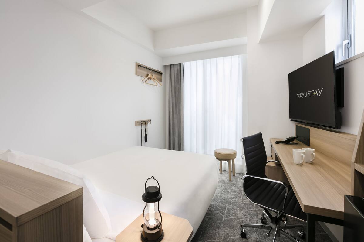 A modern hotel room with a bed, a desk with a chair, a television, and a small table with a lantern.