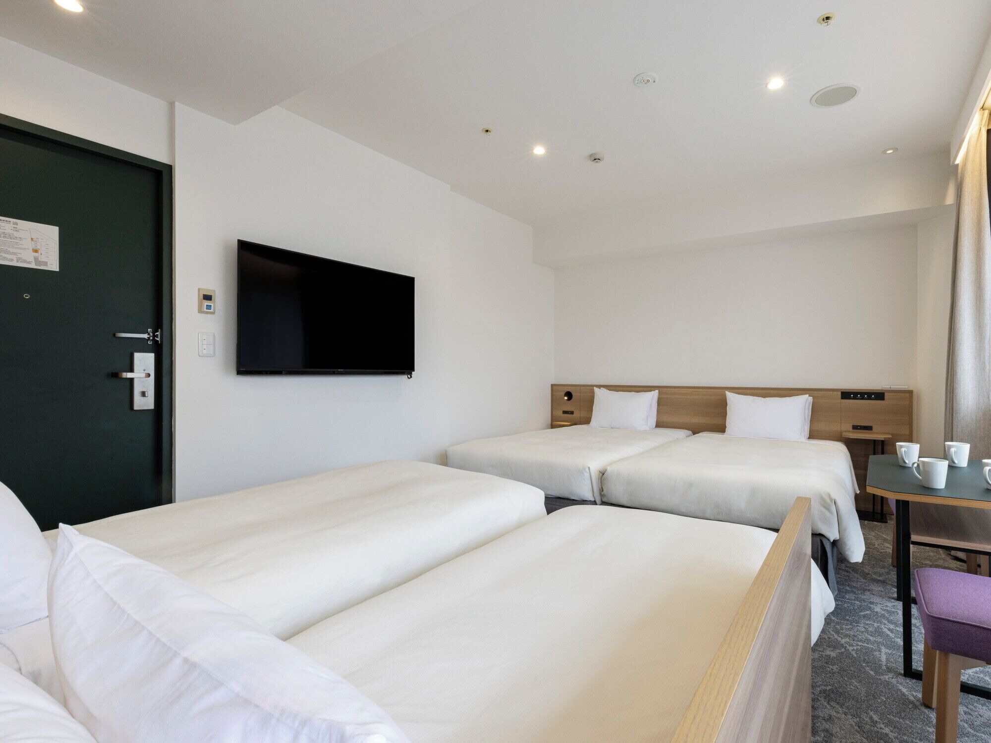 A hotel room with two single beds, a desk with a chair, a flat-screen TV mounted on the wall, and a door with a handle.