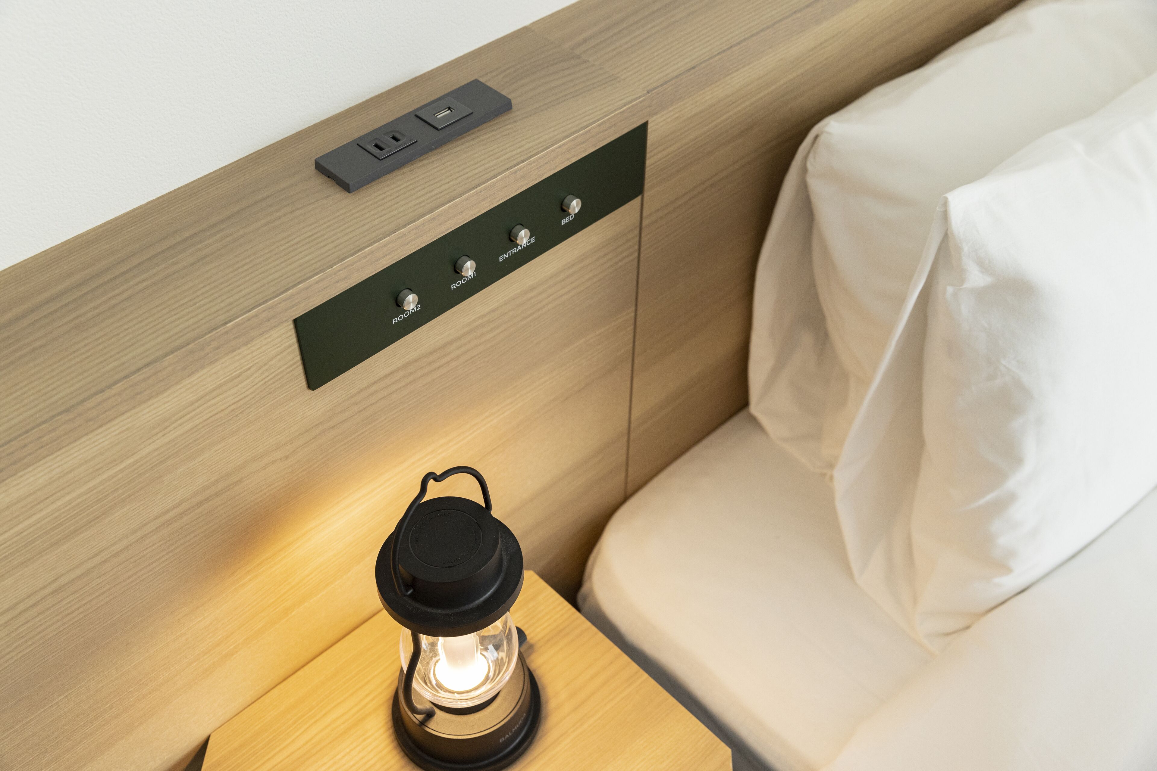 Habitación de hotel moderna con pared de paneles de madera, una mesita de noche con una linterna y un panel de control con botones de selección de la habitación.