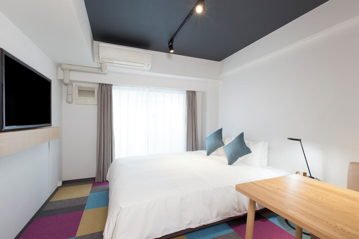 A modern hotel room with a large bed, a flat-screen TV mounted on the wall, a desk, and a window with curtains.