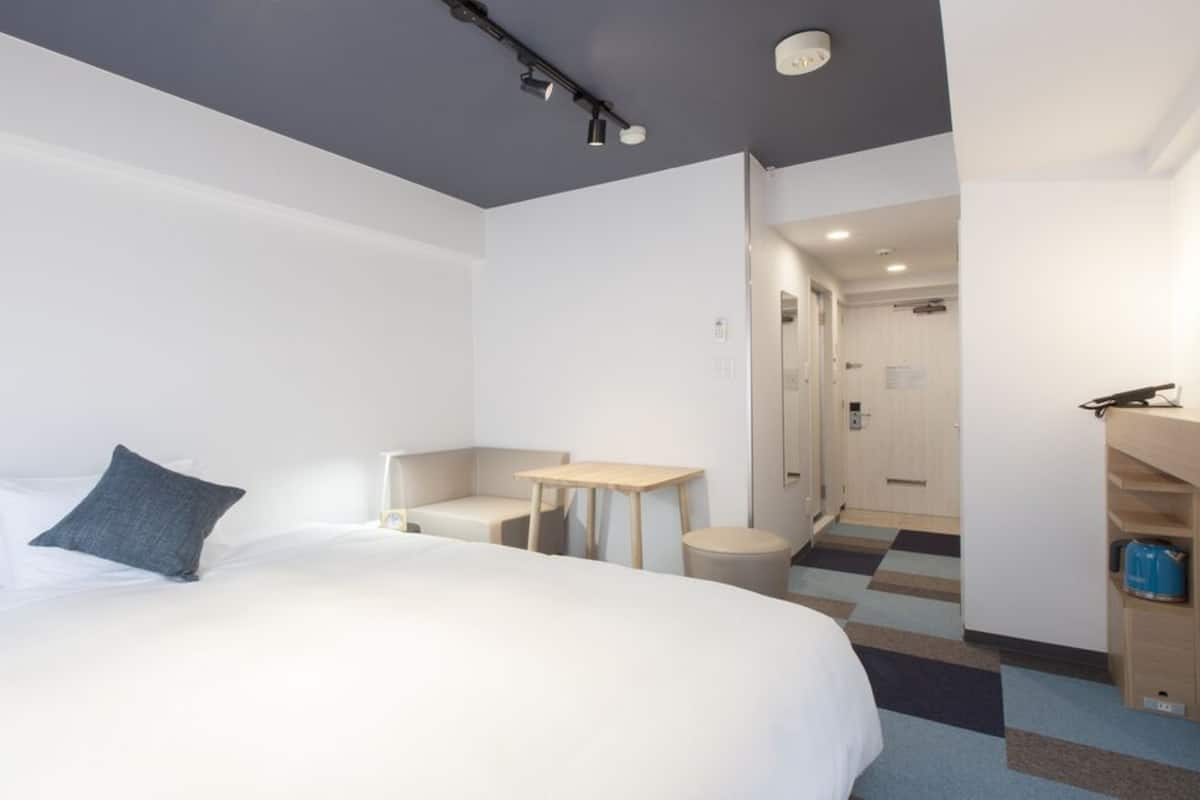 A hotel room with a bed, a desk, a chair, a small table, a trash bin, and a storage unit.