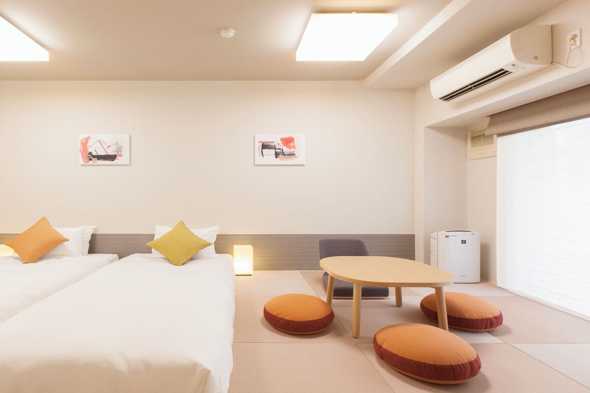 A hotel room with two beds, a small table, and a sofa. There are wall-mounted air conditioning units and a window with blinds.
