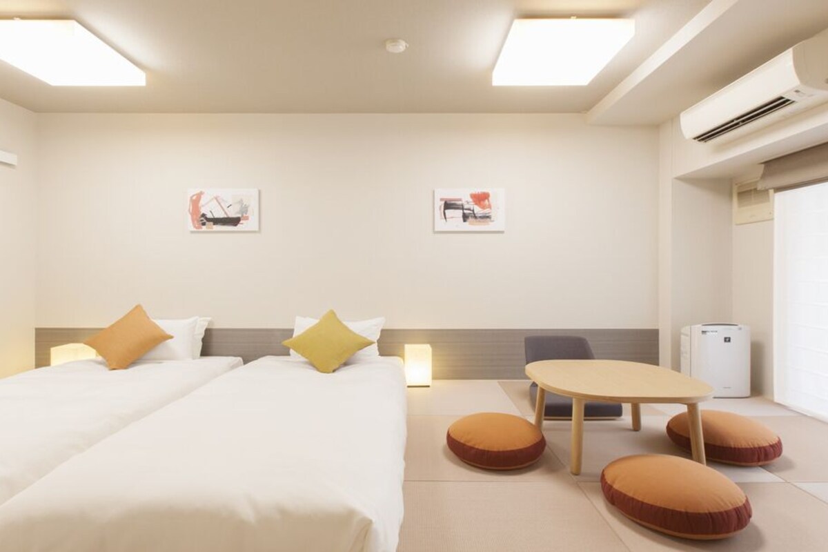 A hotel room with a bed, a table, and two chairs. There are two wall-mounted air conditioning units and a window with blinds.