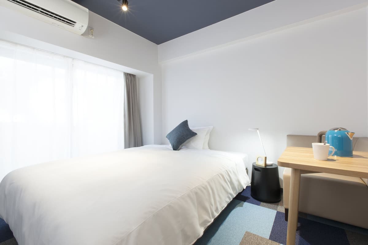 A hotel room with a bed, a desk, a lamp, a kettle, and a window with curtains.