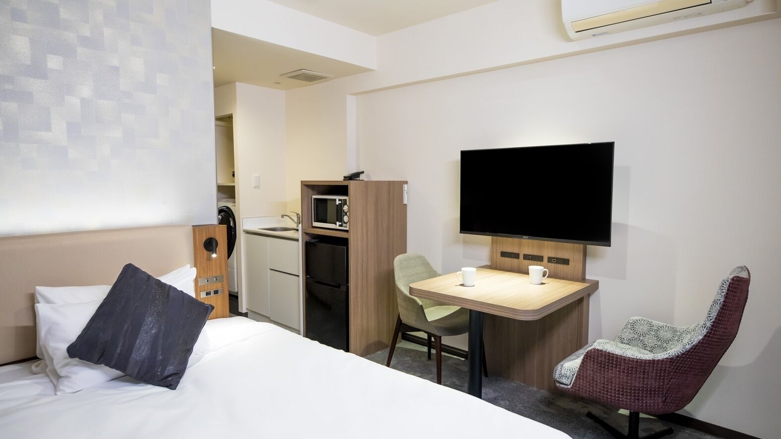 A hotel room with a bed, a desk, a chair, a TV, and a kitchenette.