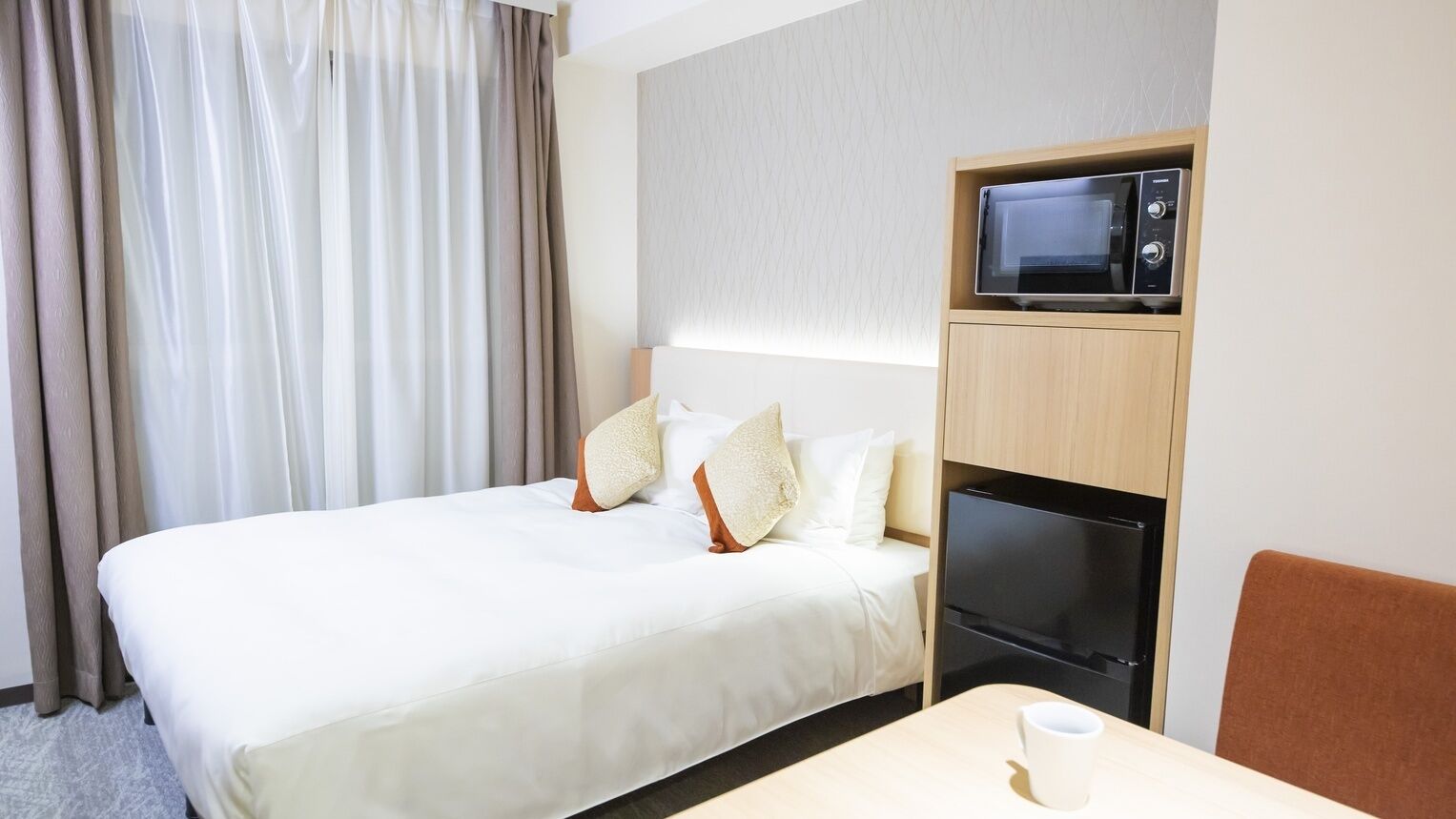 A hotel room with a bed, a microwave, a refrigerator, a table with a cup, and a chair.