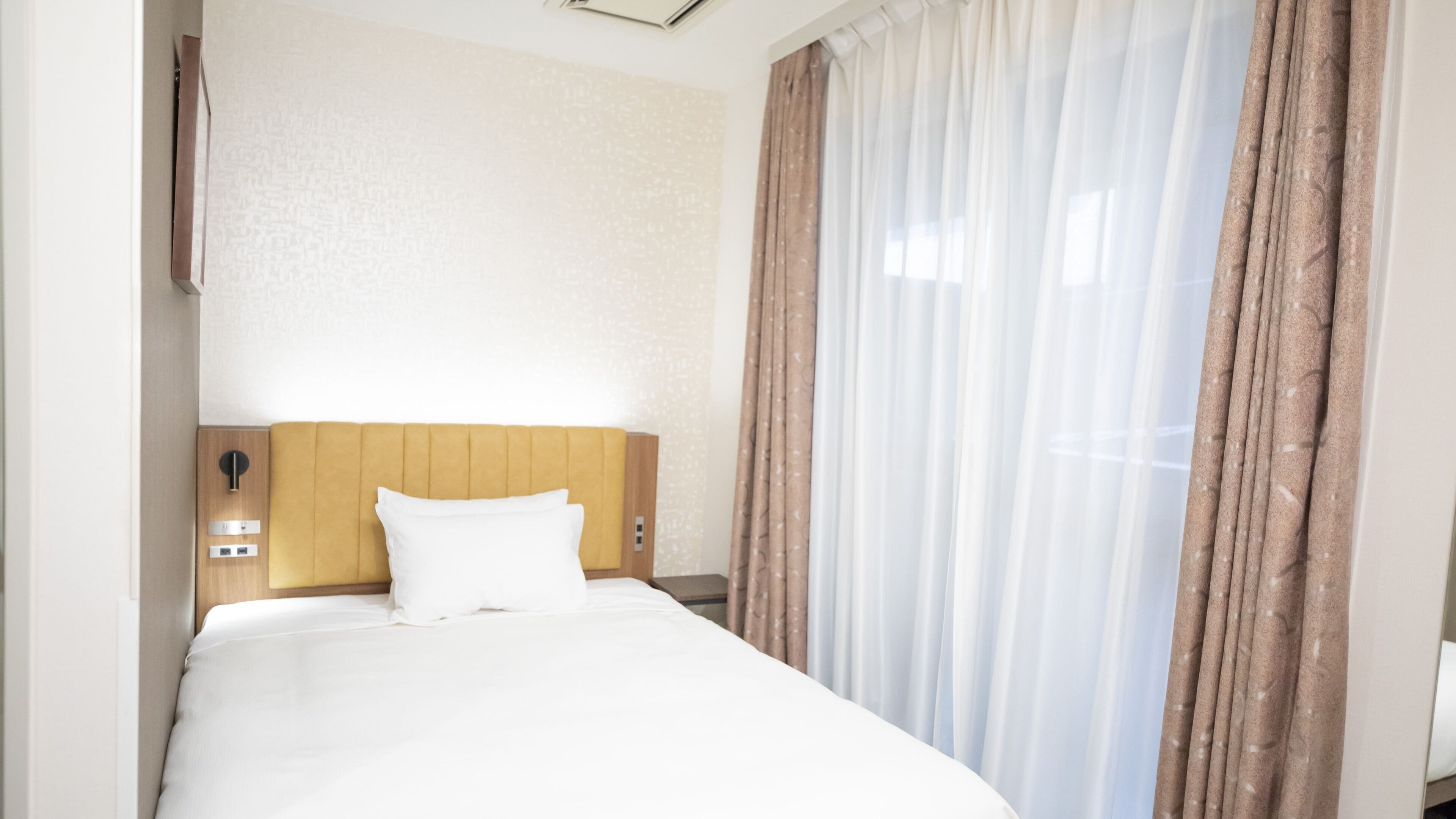 A hotel room with a bed, a headboard, a wall-mounted light, and a window with curtains.