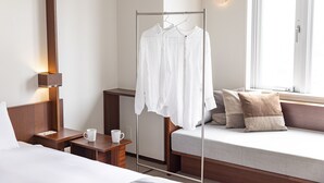 Quarto de hotel com cama, mesa de cabeceira com uma xícara, luminária, janela com vista, área de estar com almofadas e um cabideiro com camisas brancas.