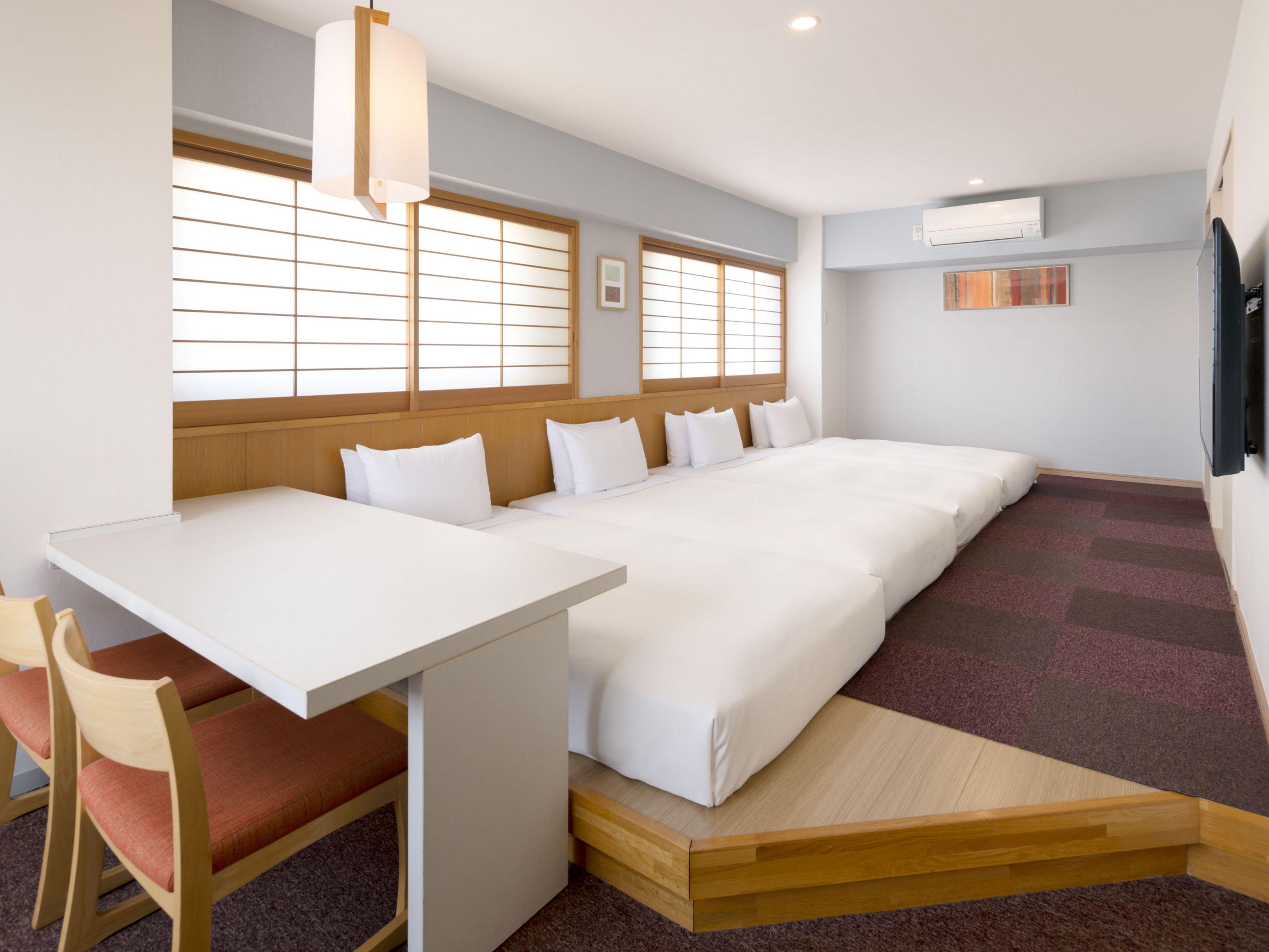 Quarto de hotel com cama, escrivaninha, cadeiras, janela com persianas e ar condicionado.