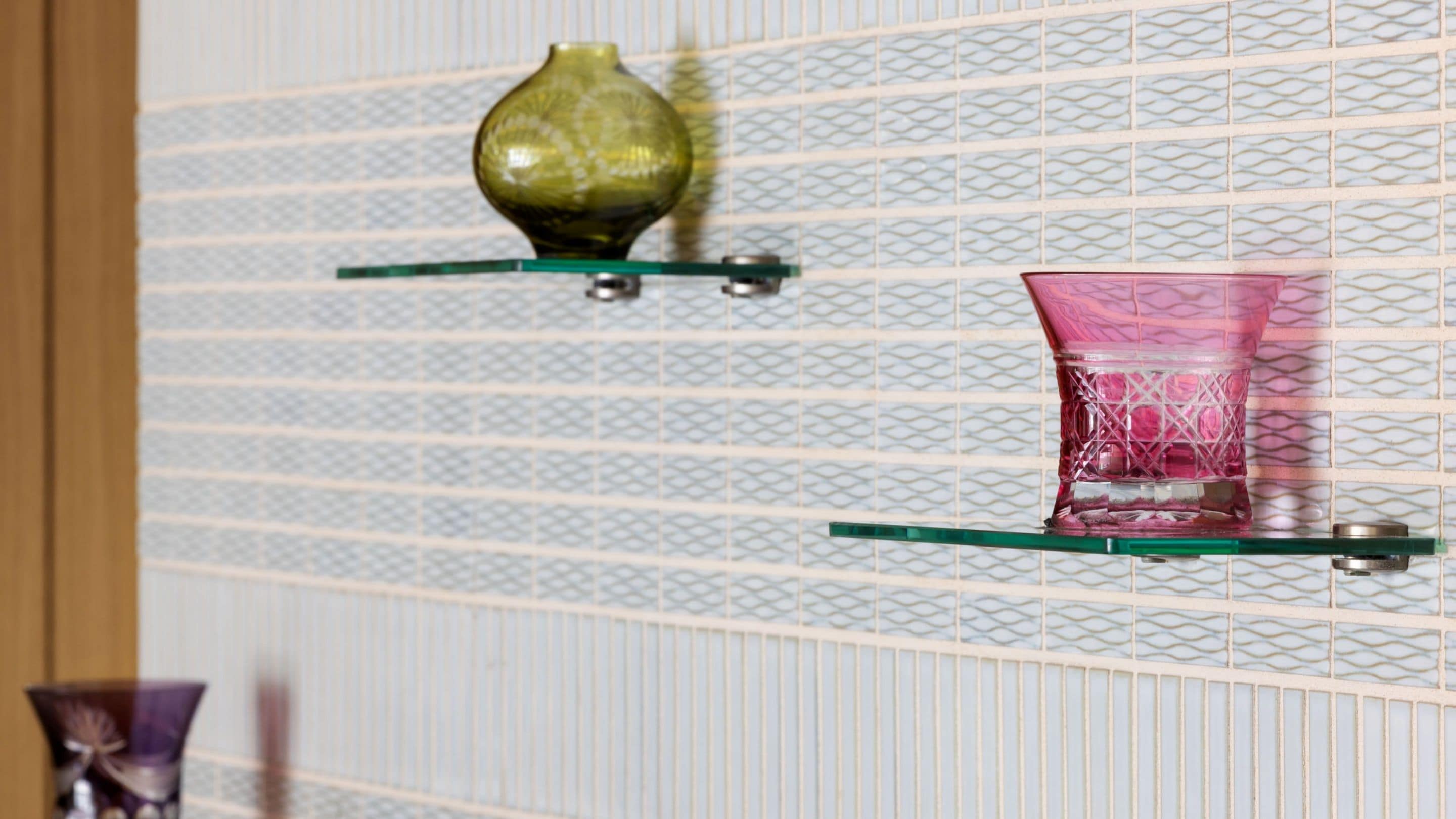 A wall with a decorative pattern and two glass shelves holding a green vase and a pink glass.