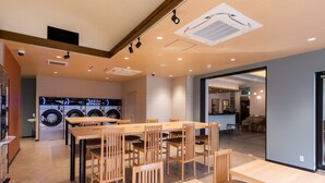 A modern laundry facility with multiple washing machines, a dining area with wooden tables and chairs, and a visible kitchen area in the background. - Tokyu Stay Monzen Nakacho Tokyo (Tokyo)