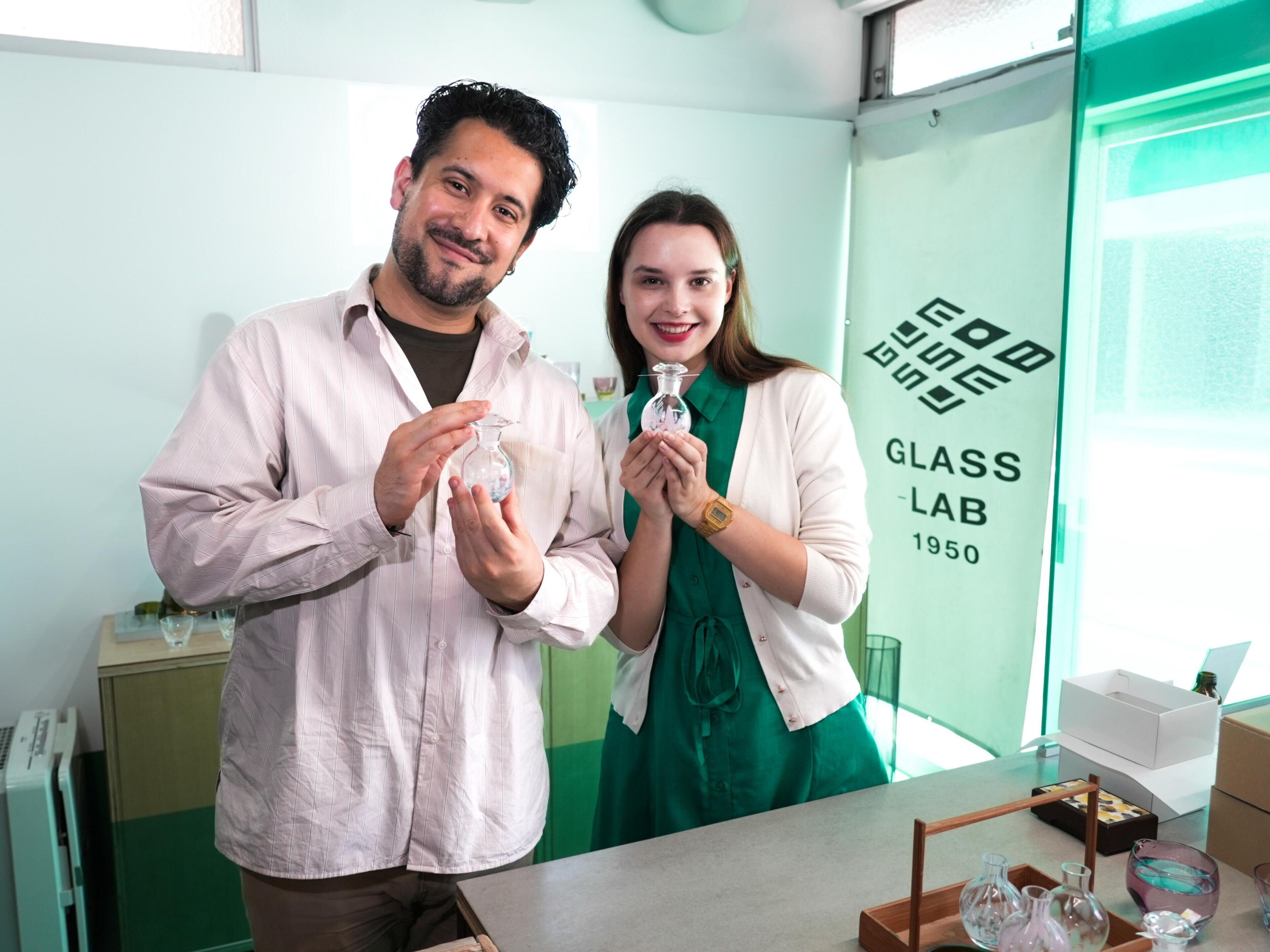 Two people in a lab setting with glassware, a green wall, and a sign reading "GLASS LAB 1950".
