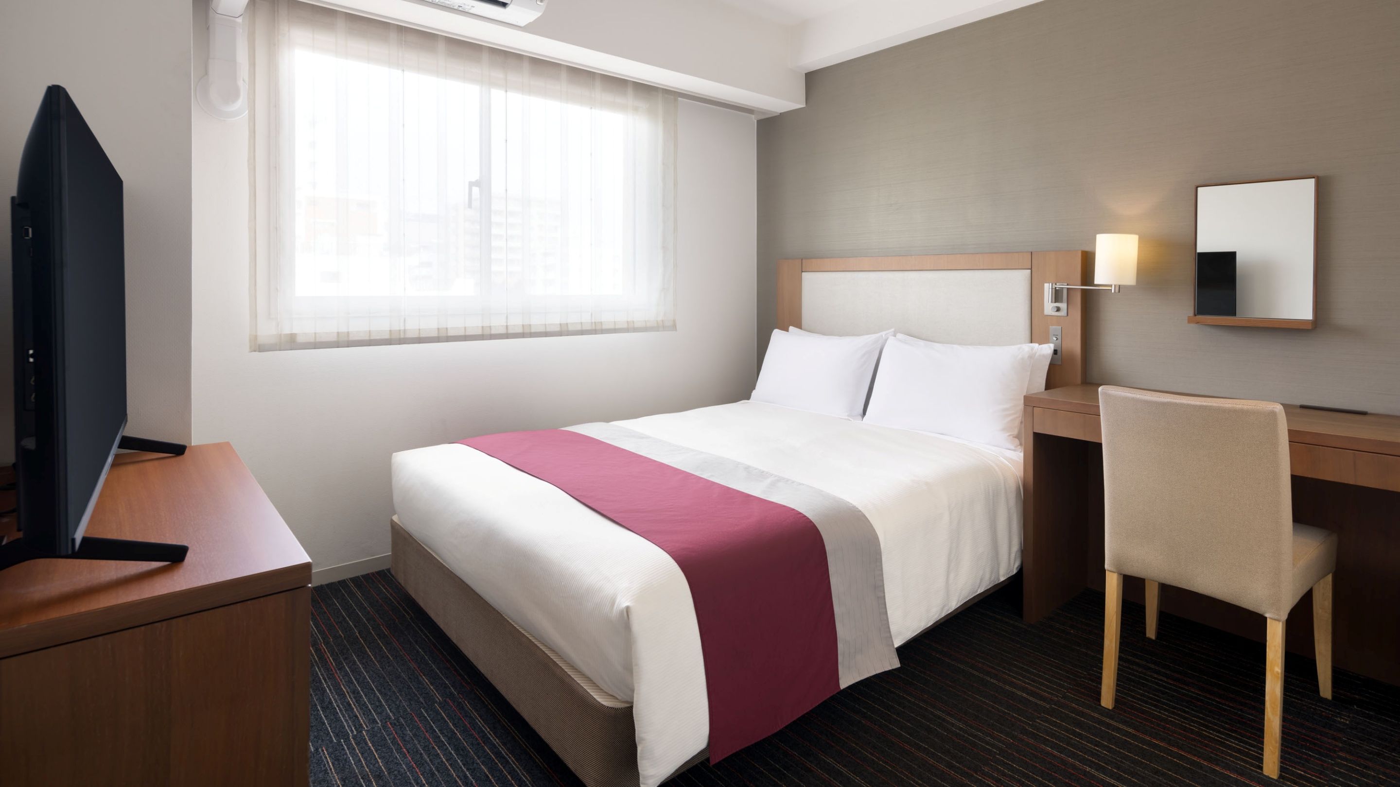 Habitación de hotel con cama, escritorio, silla, televisor y ventana con cortinas.