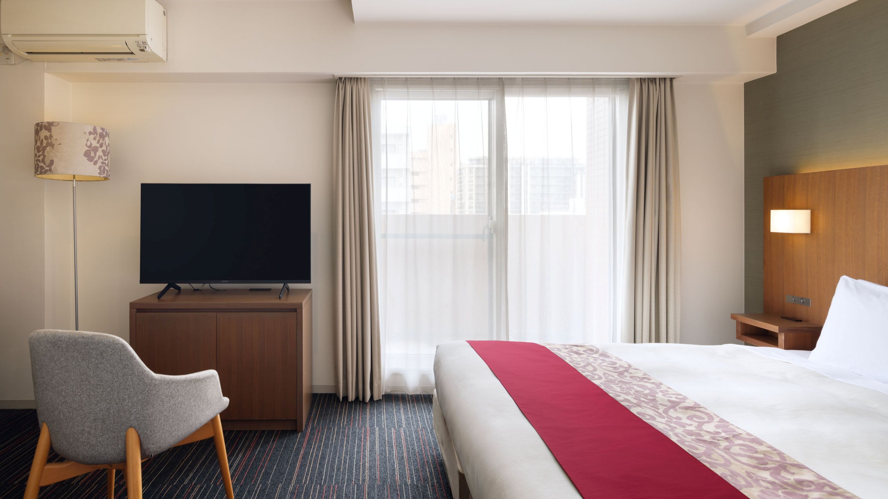 A hotel room with a bed, a television on a wooden cabinet, a desk, a chair, and a window with curtains.