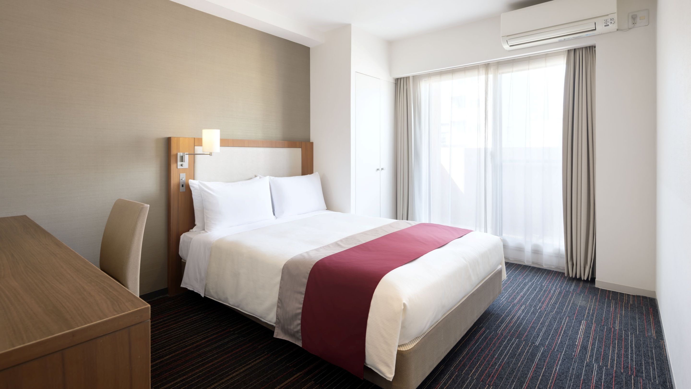 Een hotelkamer met een bed, een bureau, een stoel, een raam met gordijnen en een airconditioning.