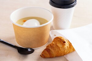A paper cup with a lid, a spoon, and a croissant on a table.