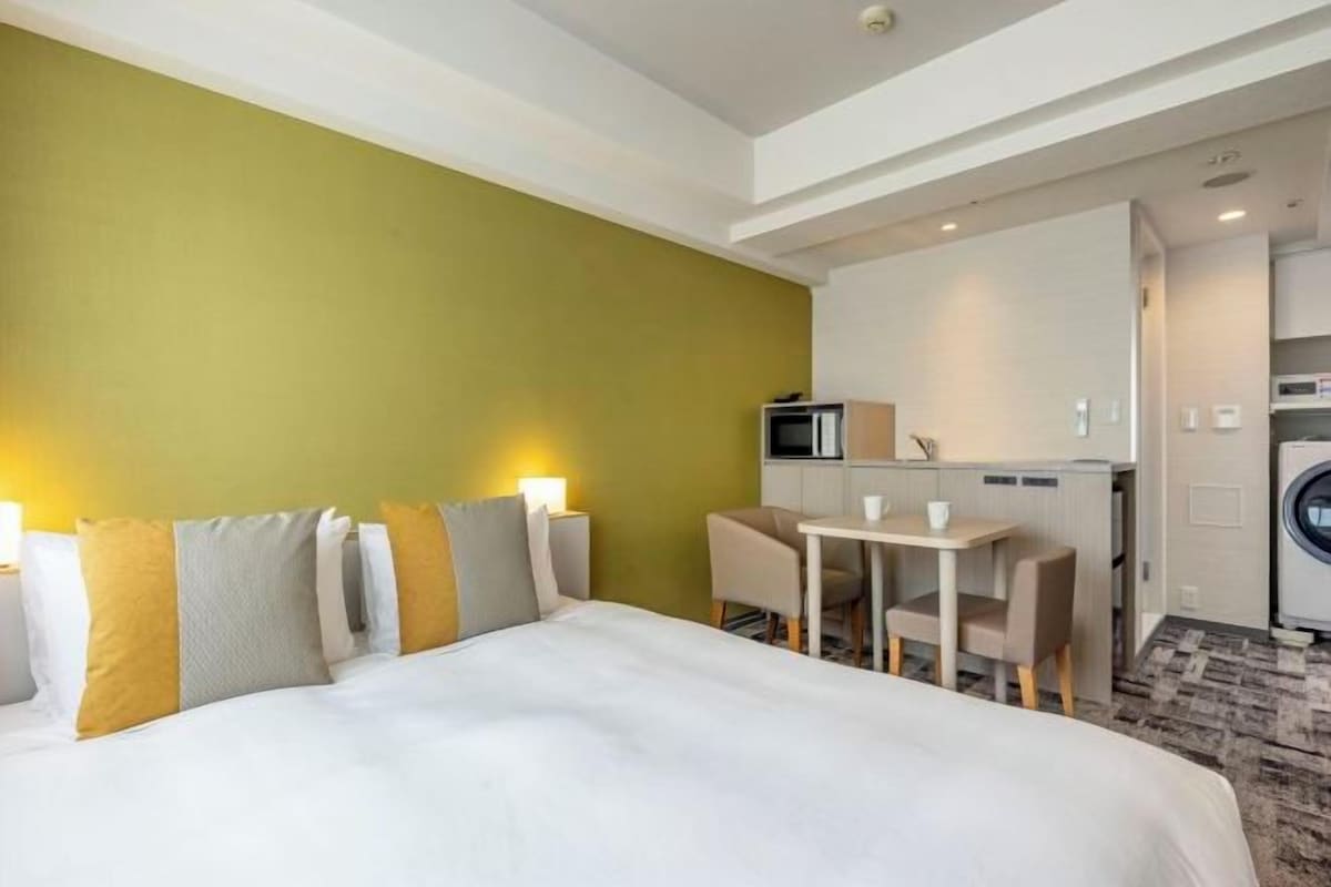 A hotel room with a bed, bedside tables, a desk, a chair, a microwave, and a washing machine.
